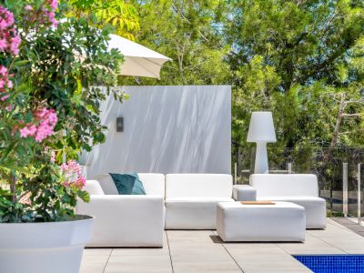 Contemporary outdoor lounge area with white modular sofas and a matching ottoman on a tiled patio, bordered by a glass railing and lush greenery. A pink flowering plant adds a touch of color.