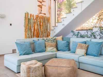 Cozy living area featuring a light blue sectional sofa, assorted cushions, and two leather poufs. A modern glass-railed staircase ascends alongside a natural wood and plant screen, with a stone wall visible beyond.