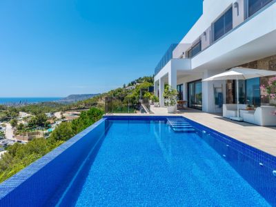 Infinity pool with blue mosaic tiles and integrated steps, bordered by a spacious terrace featuring white lounge furniture and panoramic coastal and city views.