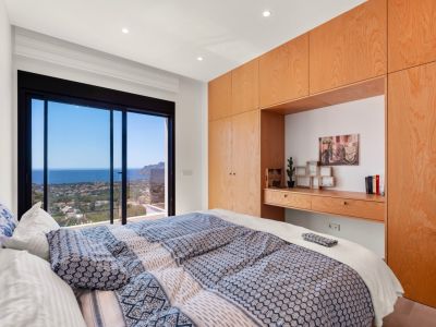 Master bedroom featuring expansive sliding glass doors offering panoramic sea and coastal views. Custom light wood cabinetry with an integrated desk provides elegant storage.