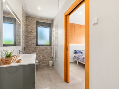 Modern bathroom featuring a grey vanity, illuminated mirror, and textured wall tiles. A frosted window and a white heated towel rail are visible, with direct access to a bedroom.