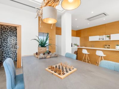 Modern dining area featuring a concrete-style table, light blue chairs, and a unique natural wood chandelier. An open kitchen with light wood paneling and a bar is visible, alongside a mosaic-tiled bathroom.