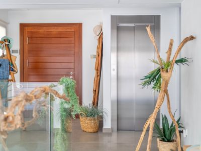 Modern interior landing featuring a sleek wooden door and a stainless steel elevator. The space is elegantly decorated with natural elements like driftwood, potted plants in woven baskets, and a glass railing.