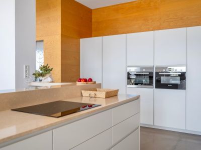 Modern kitchen featuring a sand-toned countertop with integrated induction hob. White handleless cabinets house two built-in Bosch ovens and a striking wooden accent wall.