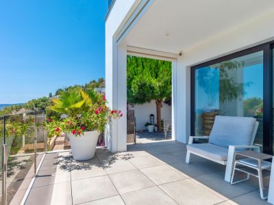 Modern outdoor terrace with light grey tiled flooring and a glass railing, offering a clear view of the verdant hillside. Features comfortable outdoor seating and vibrant potted plants under bright natural light.