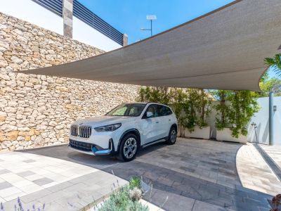 Outdoor parking area featuring a large shade sail over a patterned paved driveway. A natural stone wall and green planters define the space, with a white SUV parked.