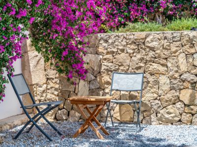 Outdoor seating area featuring two grey folding chairs and a wooden table on gravel. A natural stone wall is adorned with vibrant magenta bougainvillea.