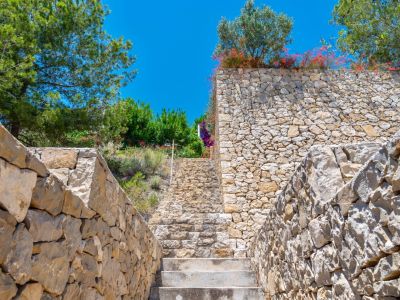 Outdoor stone staircase ascending between dry-stacked natural stone walls, surrounded by lush Mediterranean trees and flowering shrubs under a clear blue sky.