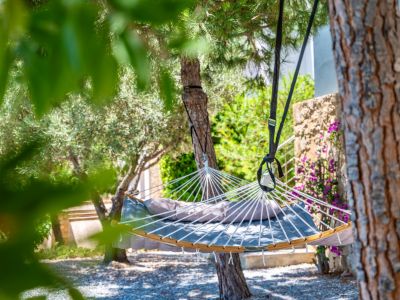 Padded grey hammock with a cushion, suspended between lush trees on a gravel ground. A stone wall with purple flowers adorns the garden background.