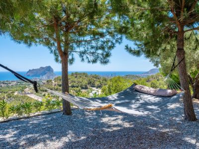 Padded hammock suspended between pine trees, providing a serene spot with panoramic views of the Mediterranean Sea and the prominent Peñon de Ifach. Gravel ground.