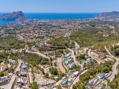 Panoramic aerial view of Calpe, showcasing the Peñon de Ifach, the coastline, and a residential area with villas and pools amidst green hills and winding roads.