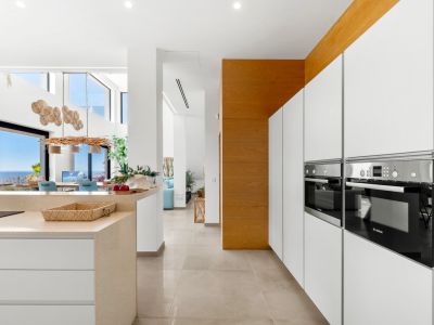 Sleek modern kitchen with integrated Bosch ovens, white handleless cabinetry, and a light stone island. The open layout extends to a dining area with panoramic sea views through large glass doors.