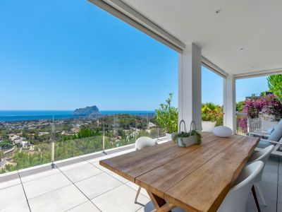 Spacious covered terrace featuring a solid wood dining table, white modern chairs, and light grey tiles. Offers panoramic sea views of Calpe and the iconic Peñon de Ifach through a clear glass railing.