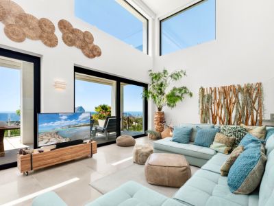 Spacious living room featuring high ceilings, large glass doors, and upper windows framing panoramic sea views. A light blue sectional sofa complements the natural wood accents and unique wall decor.