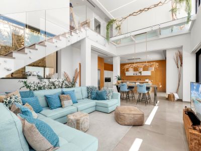 Spacious open-plan living area featuring a light blue sectional, a modern glass-railed staircase, and a kitchen with warm wood accents, all bathed in natural light.