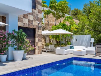 Spacious terrace featuring a blue mosaic pool and white outdoor furniture under umbrellas, complemented by a stone facade and lush Mediterranean greenery.