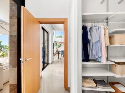 This photo captures a modern hallway entrance with an open light wood door, revealing a built-in white closet with adjustable shelves and a glimpse of a sunlit outdoor terrace with sea views.