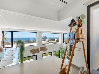 Upper level with light-toned tiled floor and glass balustrade. Expansive windows frame panoramic sea views, including Peñon de Ifach. Features a glass walkway with oval steps and a unique wooden rack.