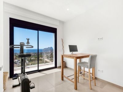 Versatile room featuring a home office setup with a wooden high desk and a fitness bike, both positioned to enjoy the expansive sea views and the iconic Peñon de Ifach through the large sliding glass doors.