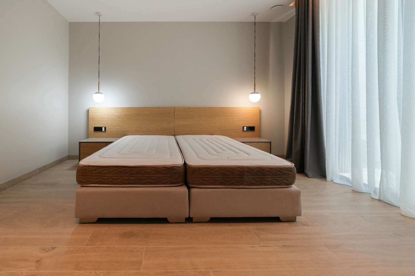 Bedroom featuring a combined double bed with upholstered bases, a light wood headboard with integrated nightstands, and two modern pendant lights. Large window with sheer curtains.