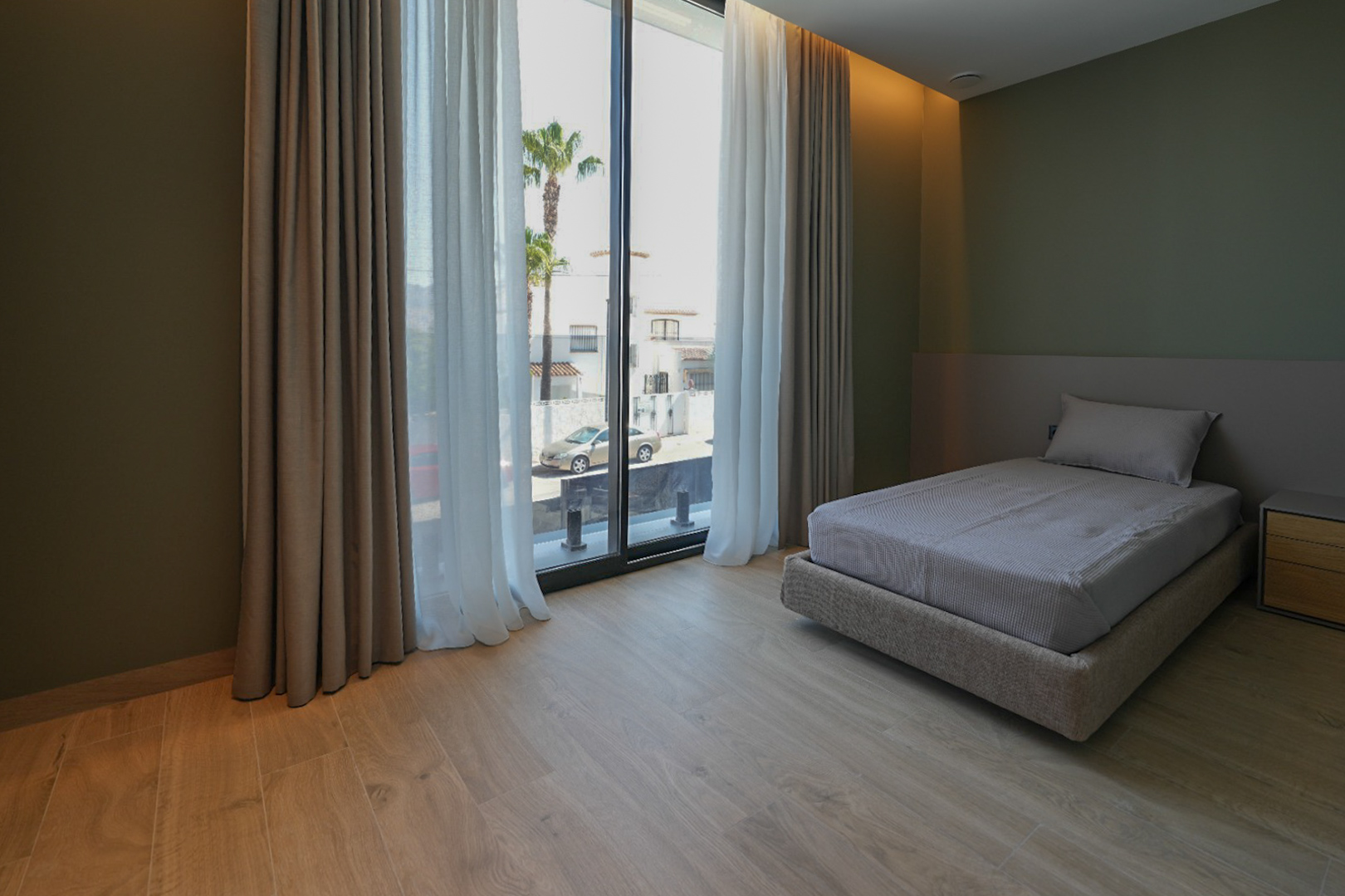 Bedroom featuring a single bed, light wood-look flooring, and olive green walls. Large windows with double curtains offer street views and palm trees.