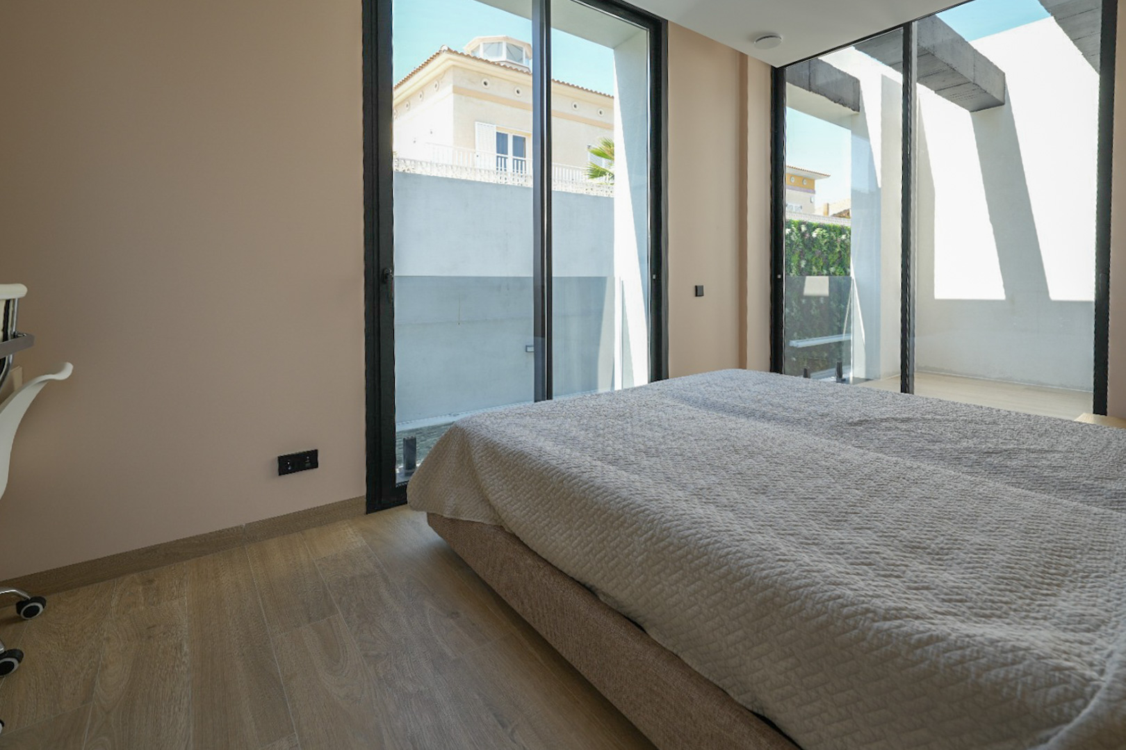 Bright bedroom featuring light wood flooring and expansive black-framed sliding glass doors, opening to an outdoor area with views of a neighboring house and green wall.