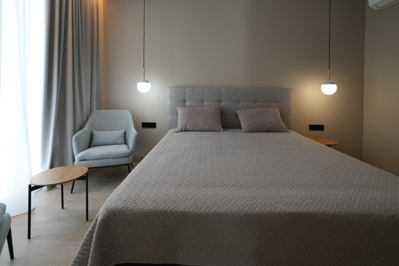 Elegant bedroom with a grey upholstered bed, two modern pendant lights, and a comfortable armchair. Light-colored curtains and wood-look flooring create a serene ambiance.