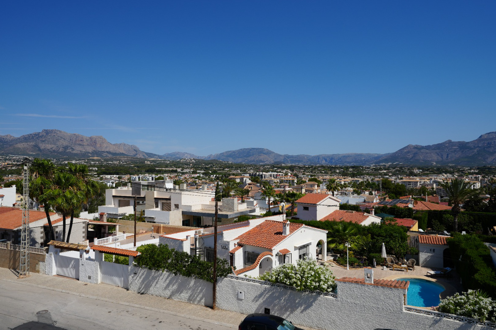 Elevated view of Albir's residential area, featuring diverse architecture, lush greenery, and a private pool, set against a backdrop of the majestic mountain range under a clear sky.