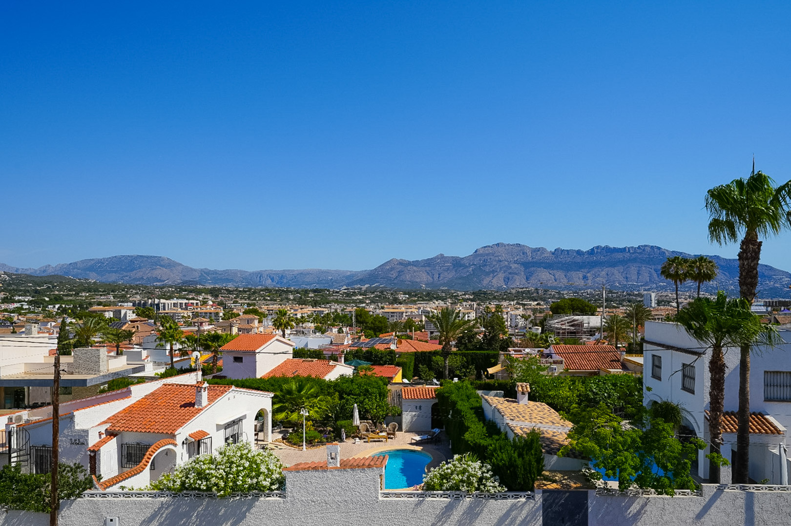 Elevated view of the Albir residential area, featuring a private pool, houses with tiled roofs, and lush Mediterranean greenery, with mountains in the background.