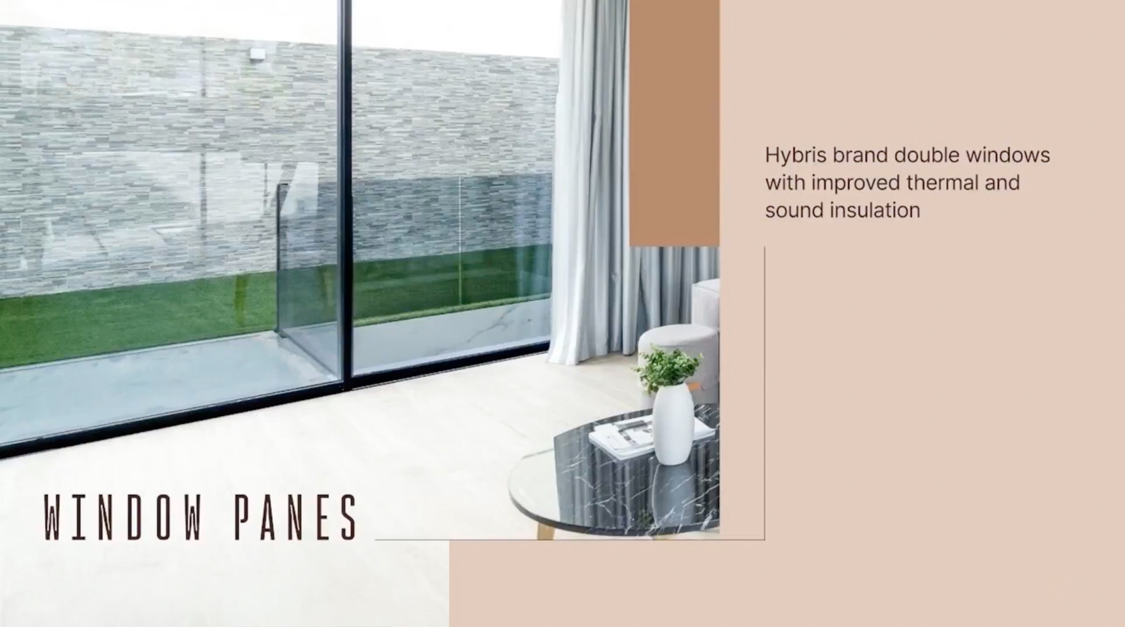 Expansive black-framed double-glazed windows overlook a textured grey stone wall and green lawn, brightening a modern interior featuring light flooring and a marble-top coffee table.