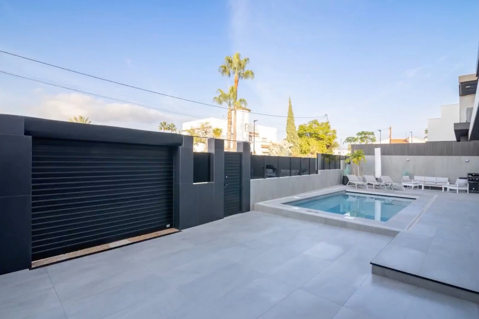 Exterior view showcasing the private pool, light grey tiled patio, and secure dark grey garage door with pedestrian gate.