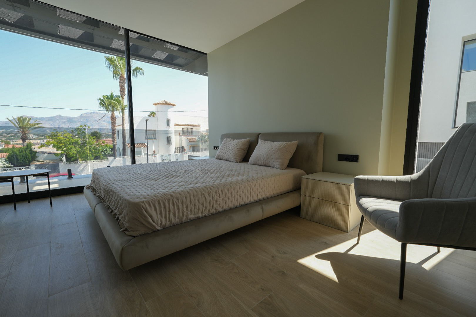 Master bedroom featuring an upholstered bed, ribbed bedside table, and expansive floor-to-ceiling windows with views of palm trees and mountains.