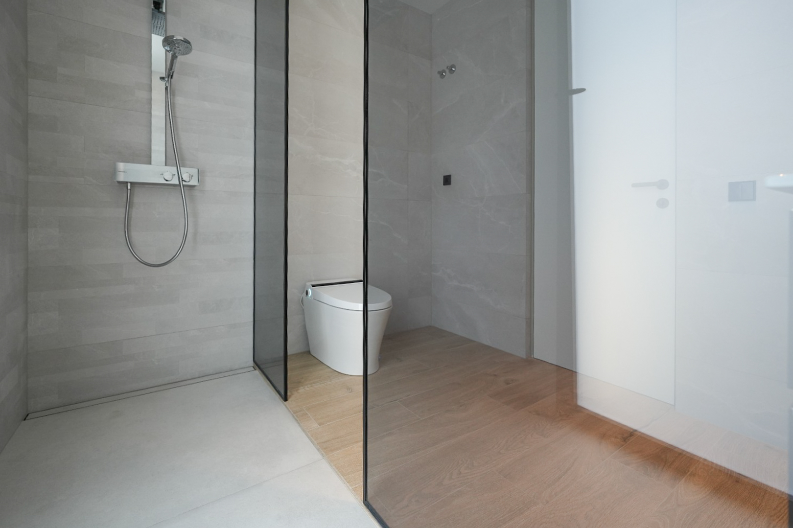 Modern bathroom featuring a walk-in shower with light grey tiles and a chrome thermostatic mixer. A glass partition separates the shower from the smart toilet area with wood-look floor tiles.