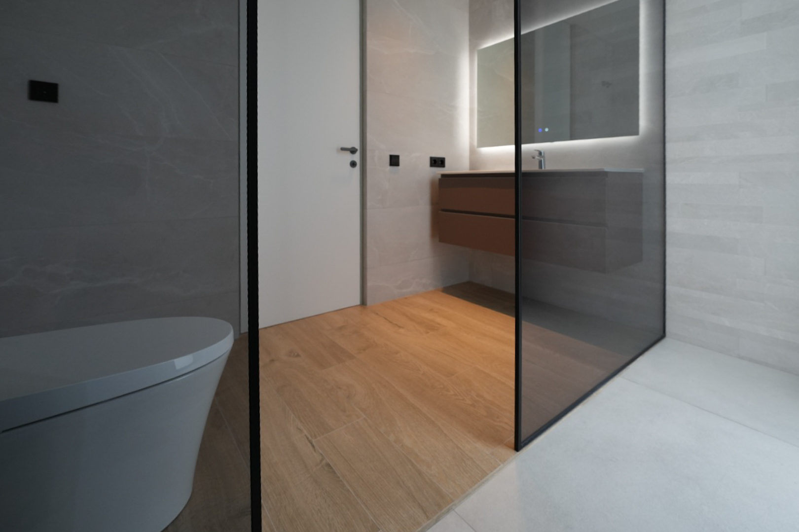 Modern bathroom featuring a wall-mounted toilet, floating wooden vanity with an illuminated mirror, and a glass shower enclosure. Wood-look floor tiles contrast with grey wall and shower tiles.