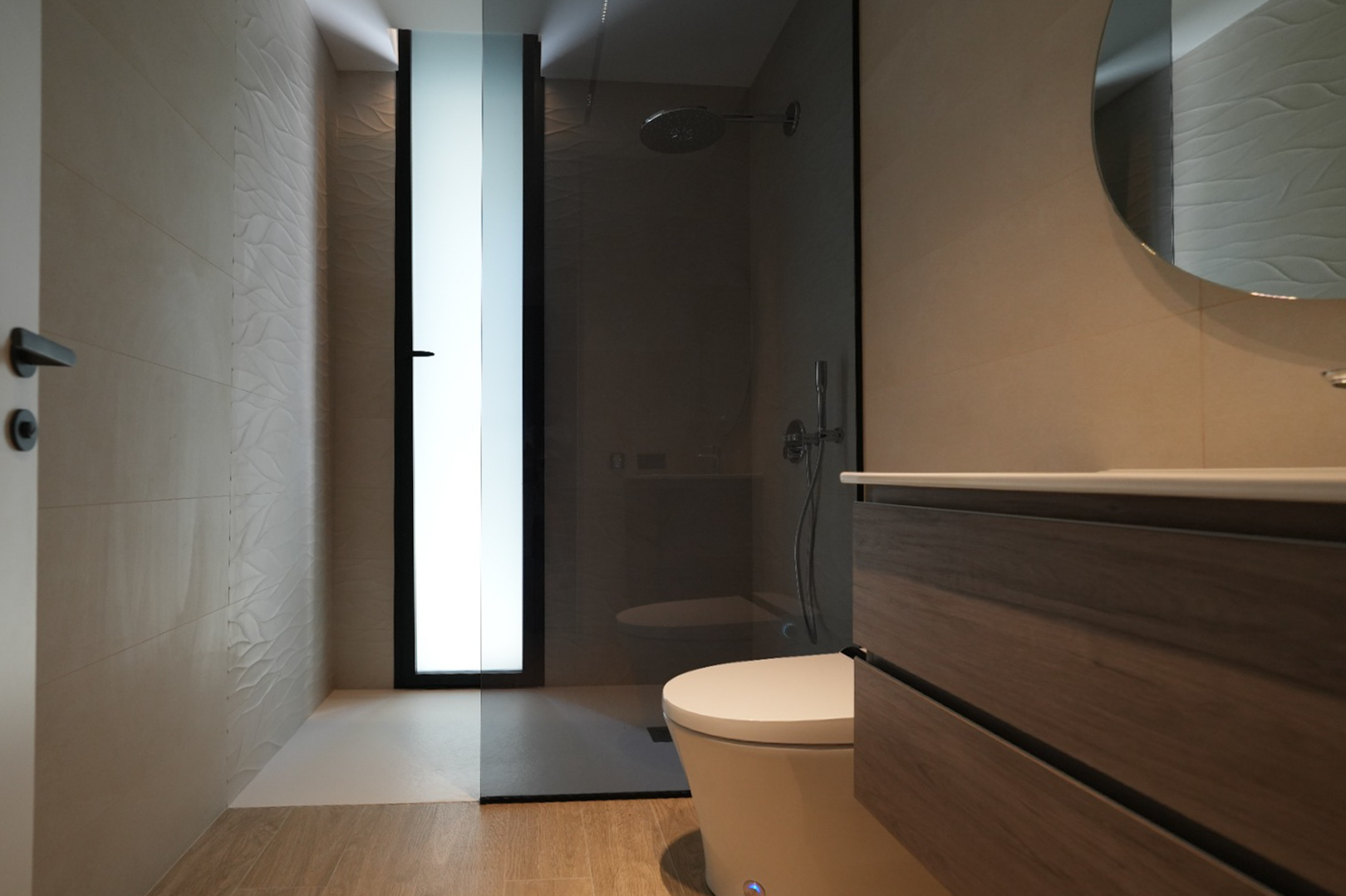 Modern bathroom featuring a wooden vanity, round mirror, and walk-in shower. Textured wall tiles and a frosted window provide light.