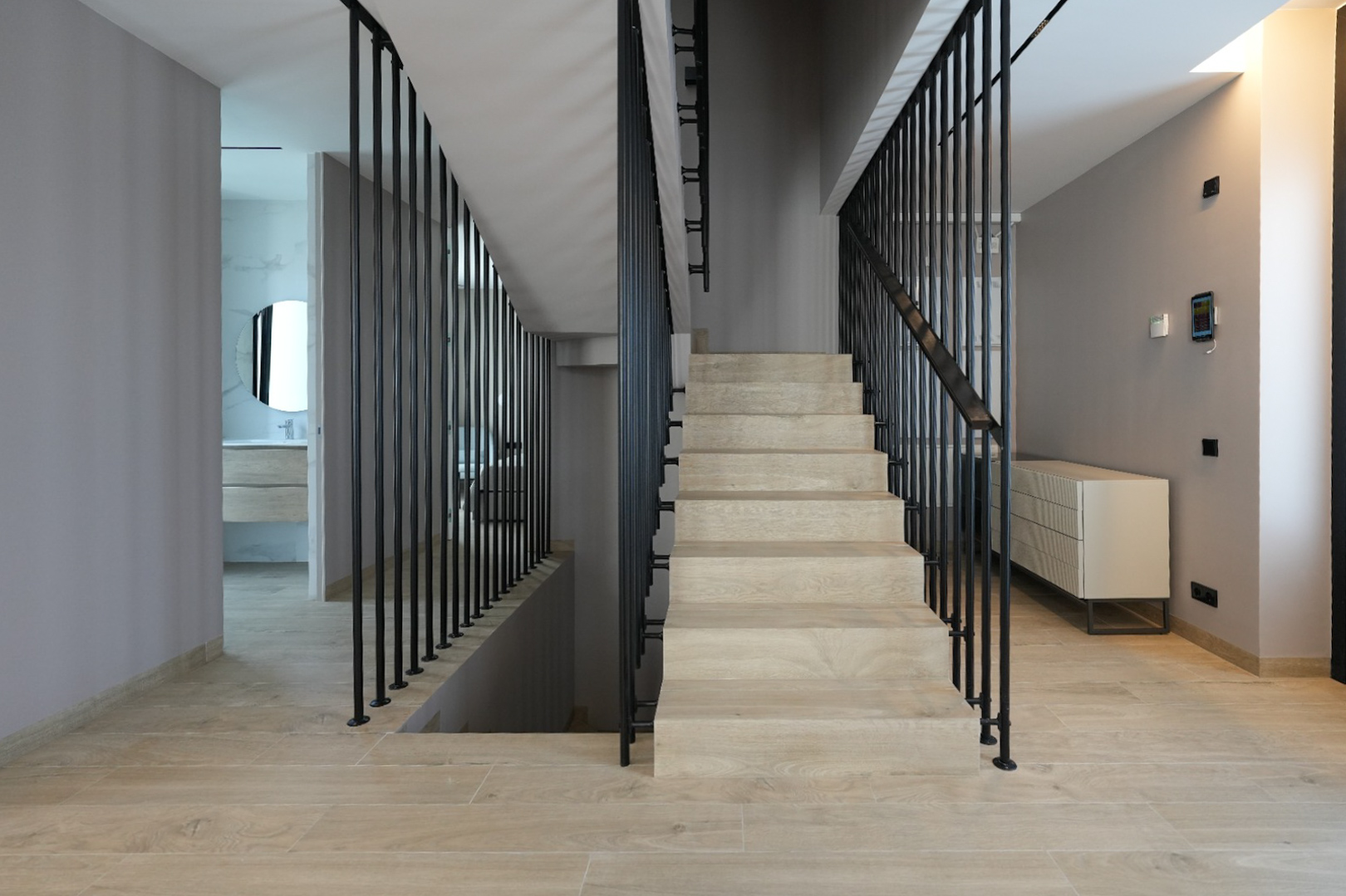 Modern interior with light wood-look staircase featuring sleek black vertical railings. A smart home control panel is visible, leading to a contemporary bathroom with a round mirror.