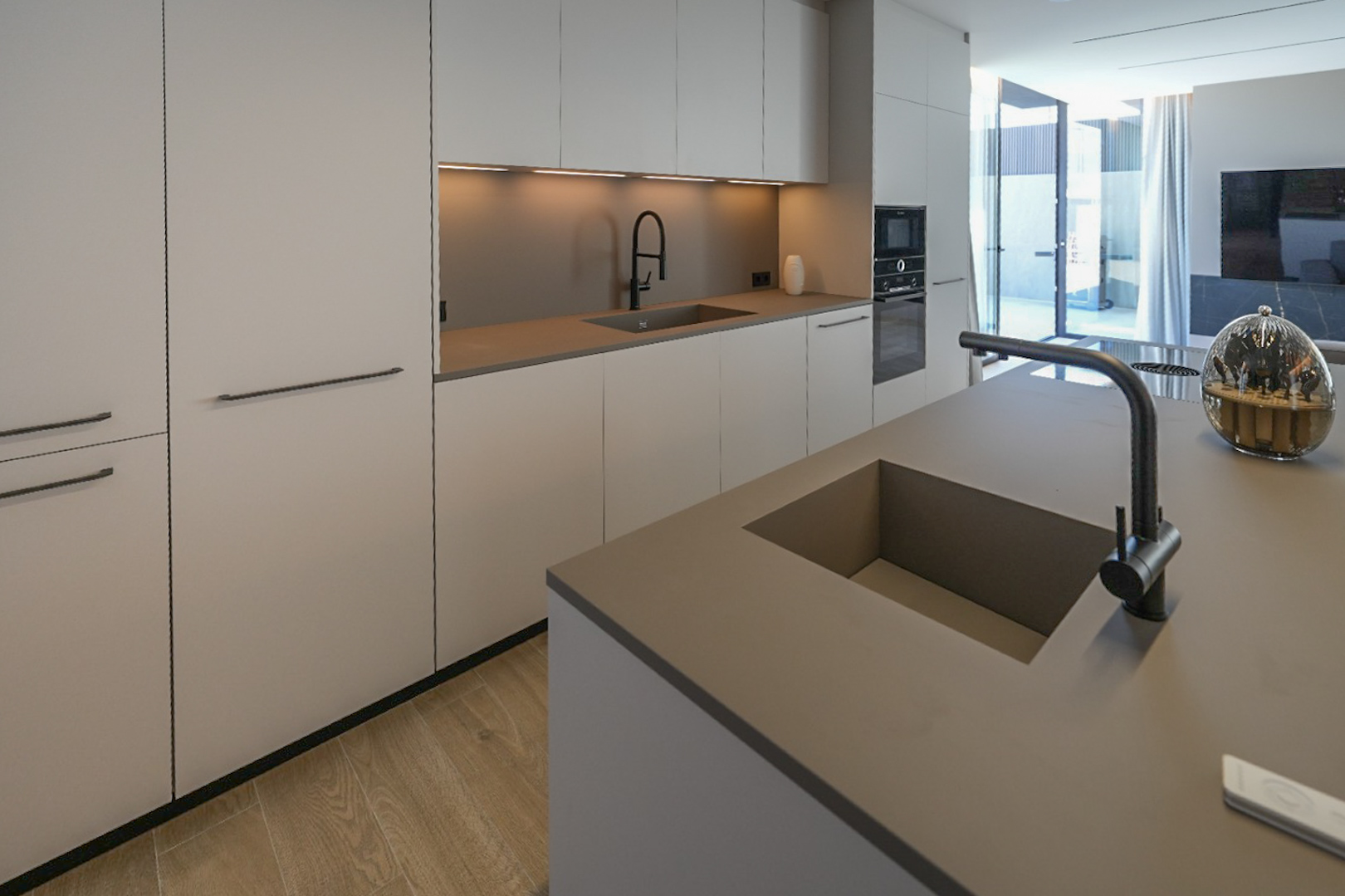 Modern kitchen featuring a central island with integrated sink, sleek flat-panel cabinetry, built-in appliances, and light wood-effect flooring, opening to an outdoor space.