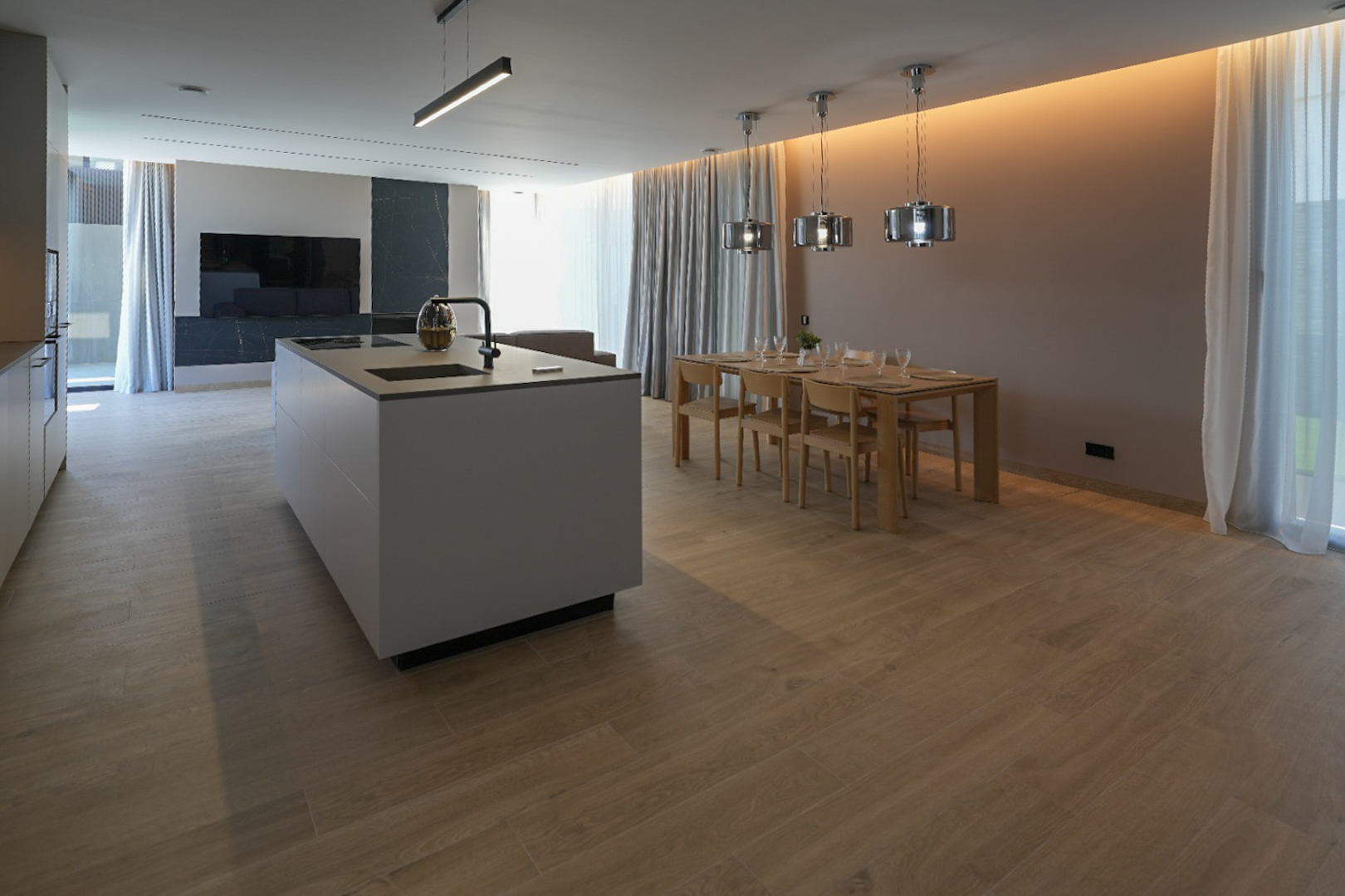 Modern kitchen island with integrated sink and hob, leading to a dining area with a wooden table, six chairs, and contemporary pendant lights. Wood-look tiled floor.