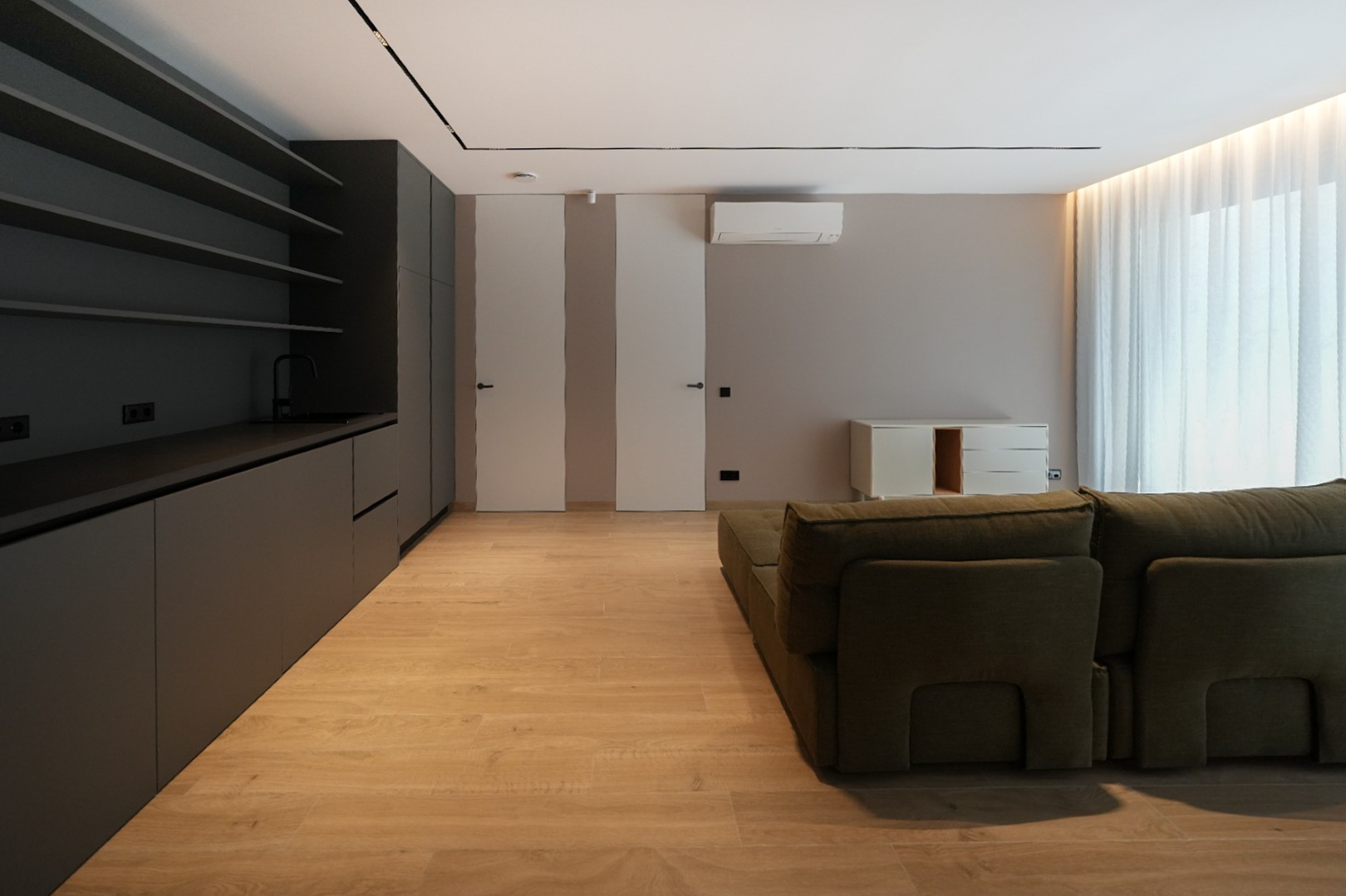 Modern living space featuring a dark grey integrated kitchenette, light wood-look flooring, and a green modular sofa. Flush doors and natural light.