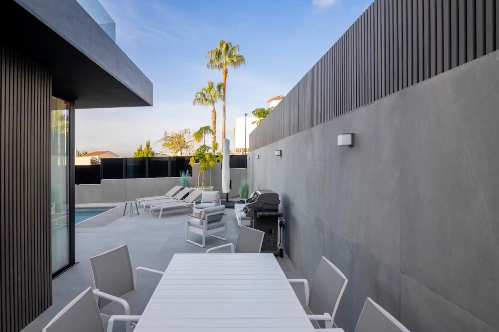 Modern outdoor terrace featuring a white slatted dining set, lounge area, and barbecue, set against grey tiled walls and dark vertical privacy screens, with a pool view.
