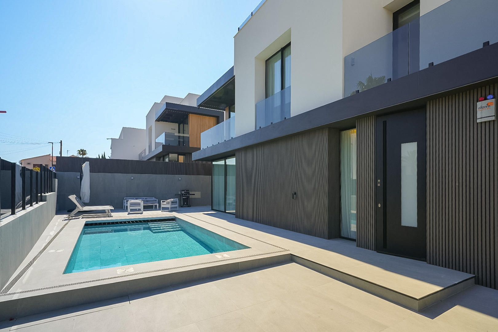 Modern villa exterior featuring a rectangular swimming pool with integrated steps, surrounded by light grey paving. The ground floor showcases a dark slatted facade and a prominent entrance door.