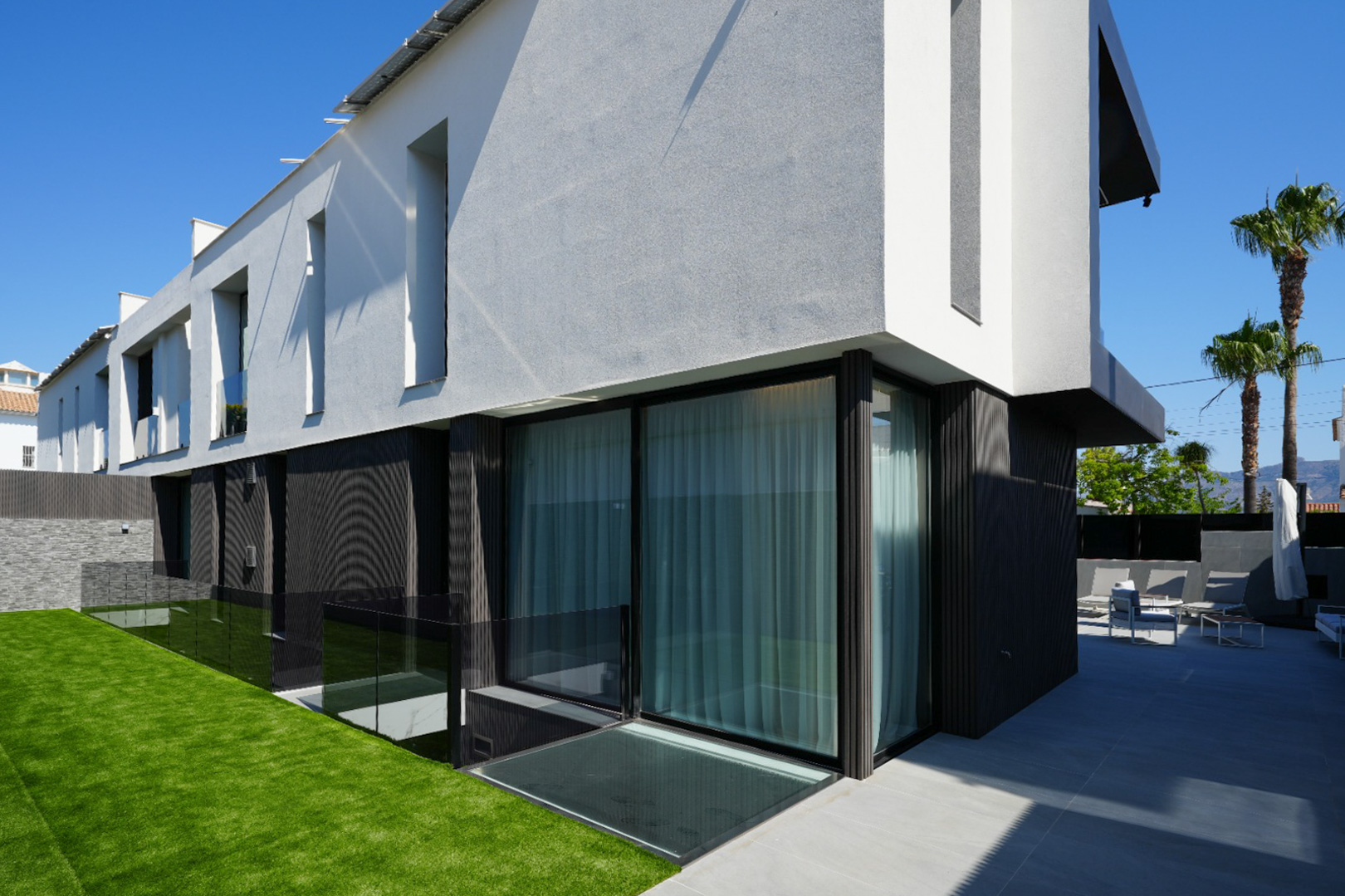 Modern villa exterior showcasing contrasting white/grey and dark textured facades, large glass doors, a manicured lawn, and a tiled patio with outdoor seating.