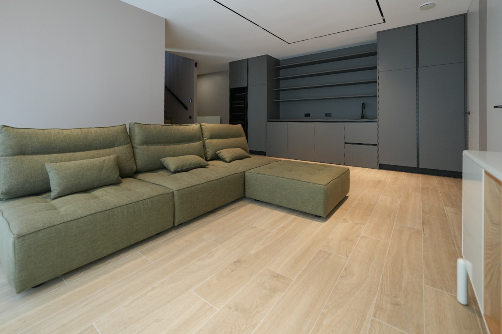 Olive green modular sofa in an open-plan living space with light wood-look tile flooring. Dark grey built-in cabinetry with a sink and shelving visible in the background.