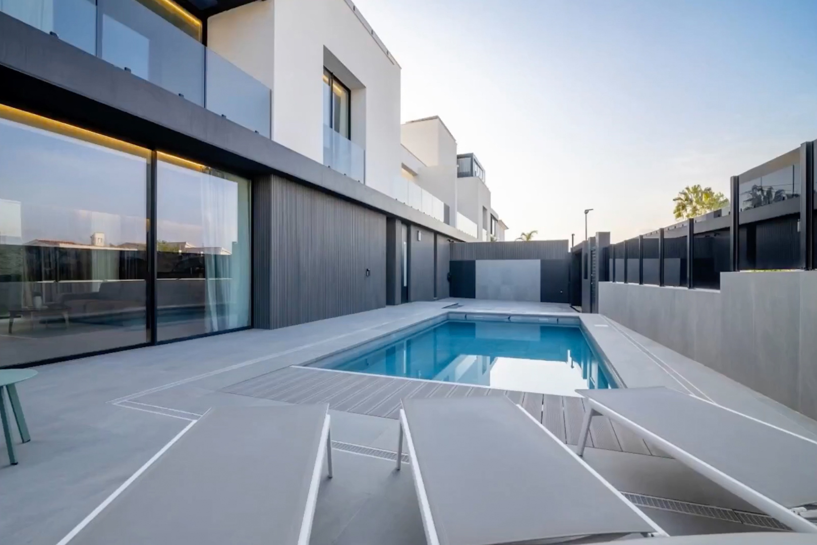 Outdoor pool with blue water, bordered by a grey tiled patio and composite decking. Modern facade with large sliding glass doors reflects the clear sky.