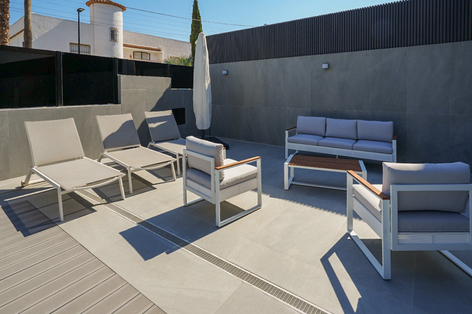 Outdoor terrace featuring modern lounge furniture in white and grey, with mixed porcelain tile and composite decking. Sun loungers and a sofa set are visible.