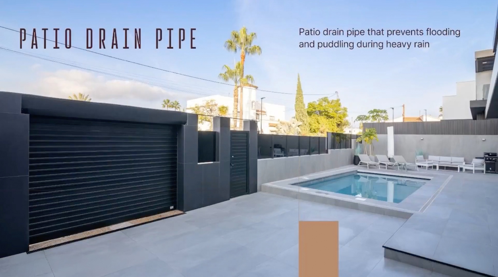 Spacious outdoor patio featuring a swimming pool, light grey tiled flooring, and a modern black garage door. Integrated patio drain pipe prevents water accumulation.