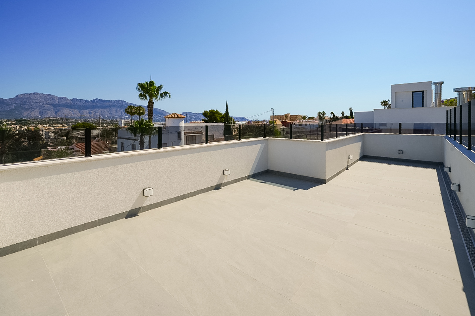 Spacious rooftop terrace with light-toned floor tiles and modern glass railing, offering panoramic views of distant mountains and the surrounding town.