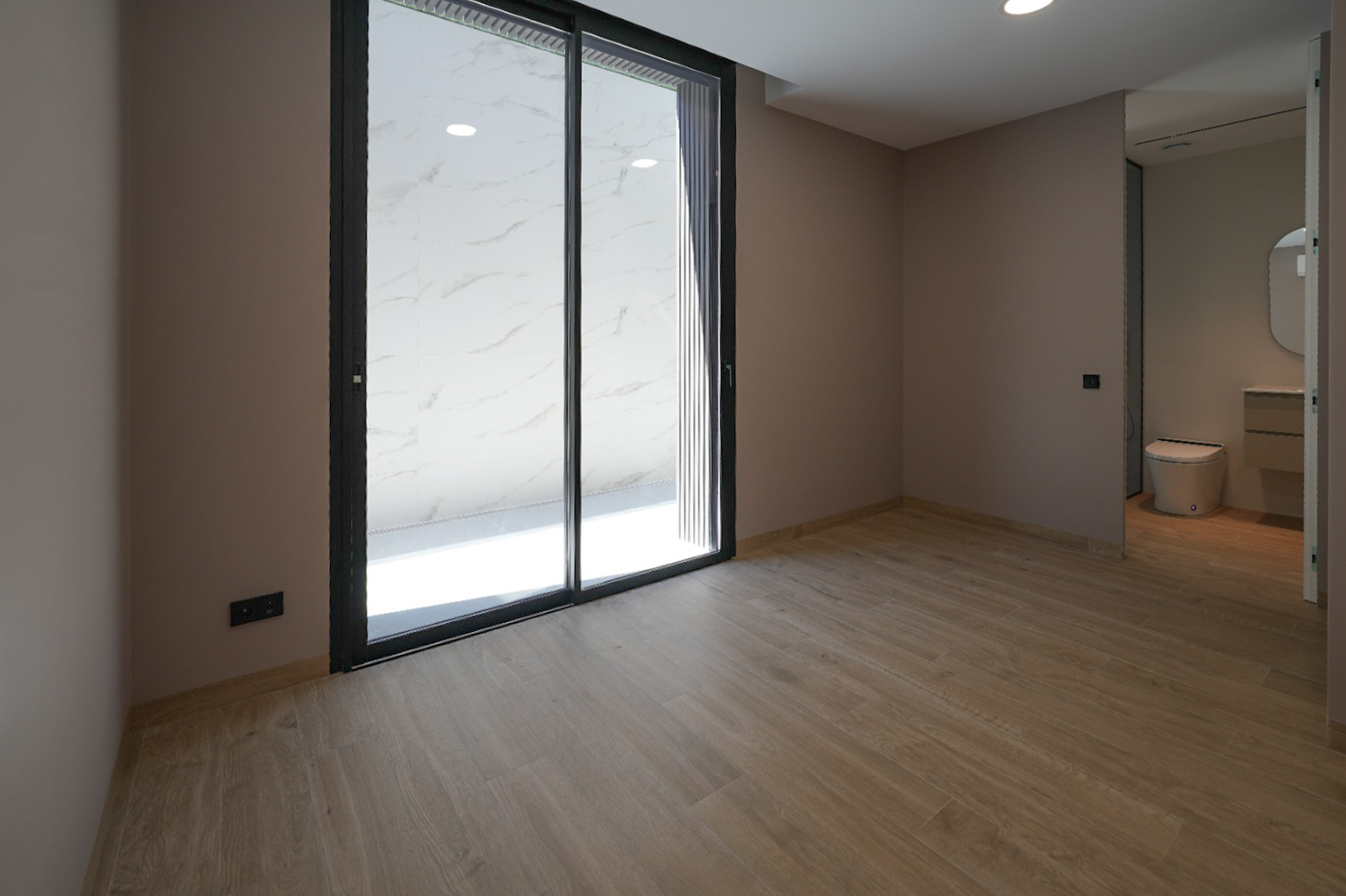 Spacious room with wood-look tiled flooring and light-colored walls. Large black-framed sliding glass doors open to an outdoor area with white marble-patterned tiles. Integrated bathroom visible.
