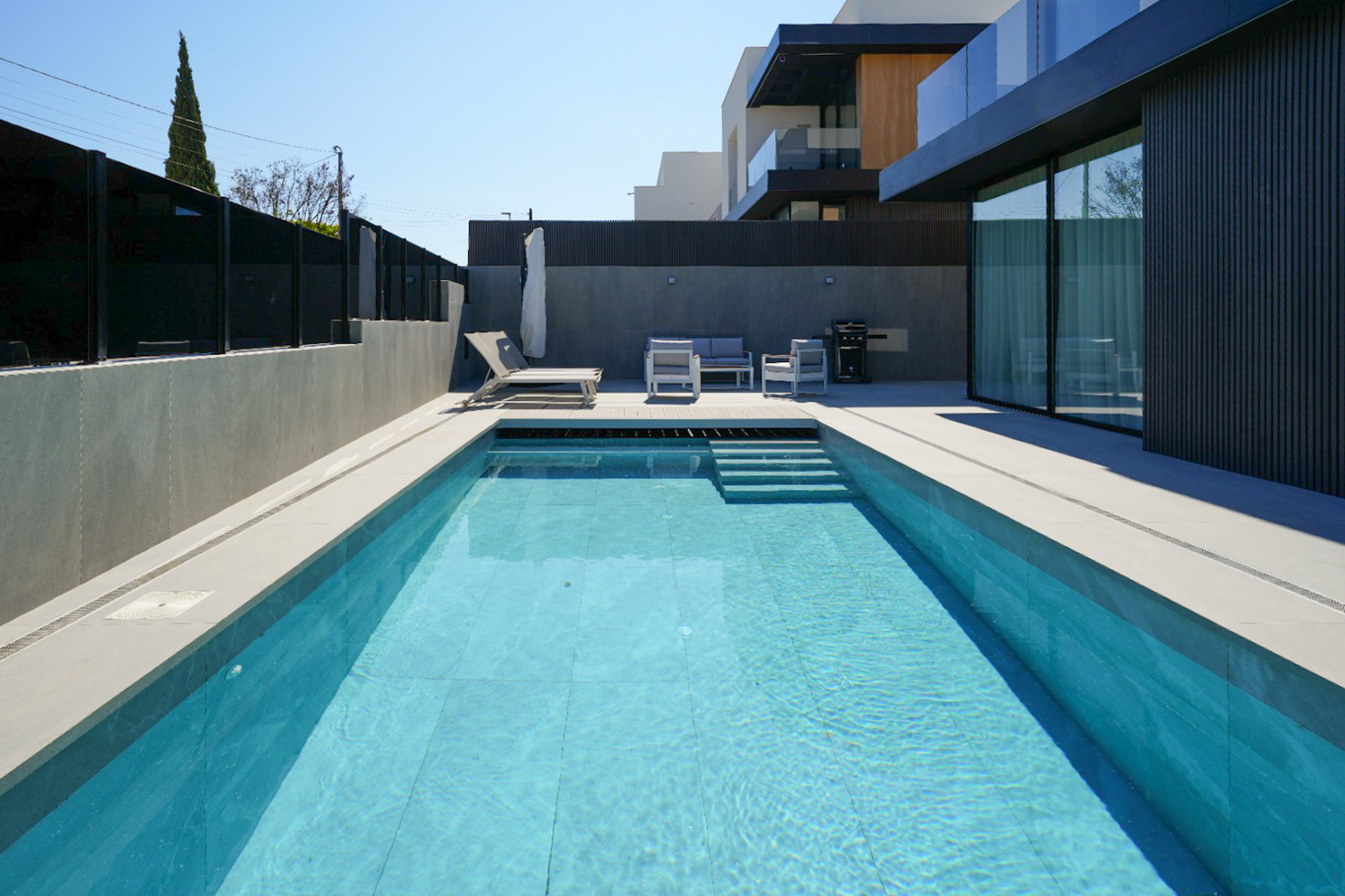 The outdoor area features a rectangular pool with light blue tiles and steps, surrounded by light grey paving. A modern lounge set and sun lounger offer relaxation beside the sleek facade.