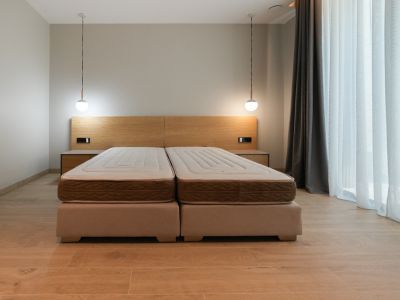 Bedroom featuring a combined double bed with upholstered bases, a light wood headboard with integrated nightstands, and two modern pendant lights. Large window with sheer curtains.